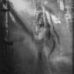 Autoportrait nu bougé, devant un mur d'une cellule des prisons militaires désaffectées de Labry en Lorraine. Sur le mur , figure un dessin représentant une femme.